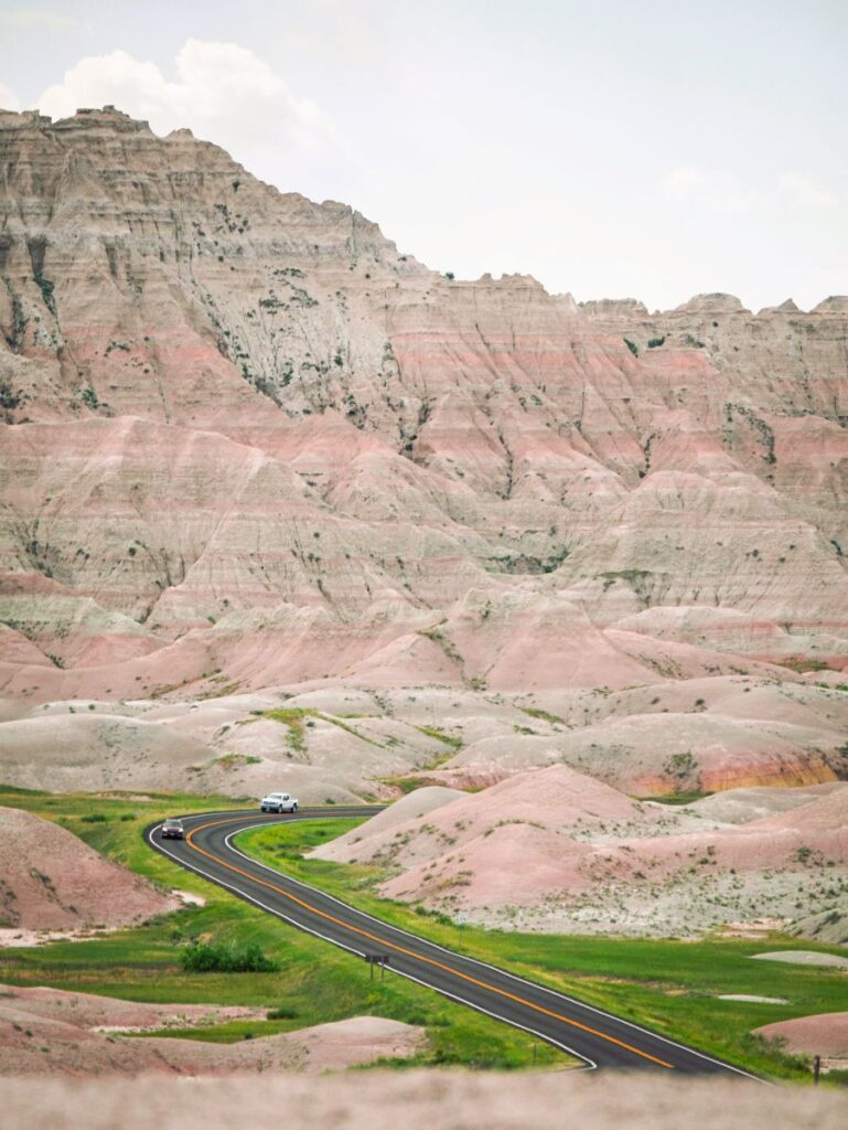 Car driving along the Badlands Loop Road surrounded by colorful layered formations and prairie.