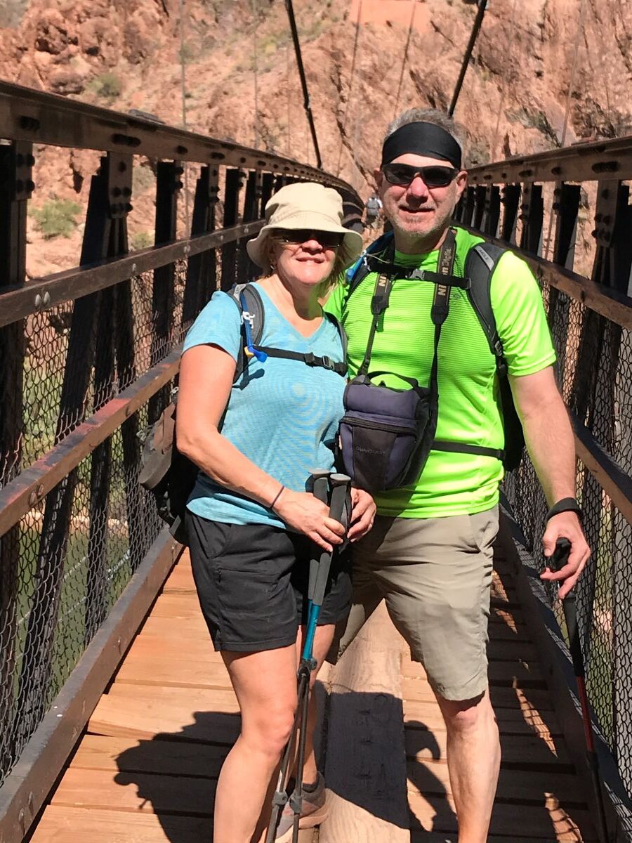 Two hikers standing on a suspension bridge in the Grand Canyon with gear and trekking poles.